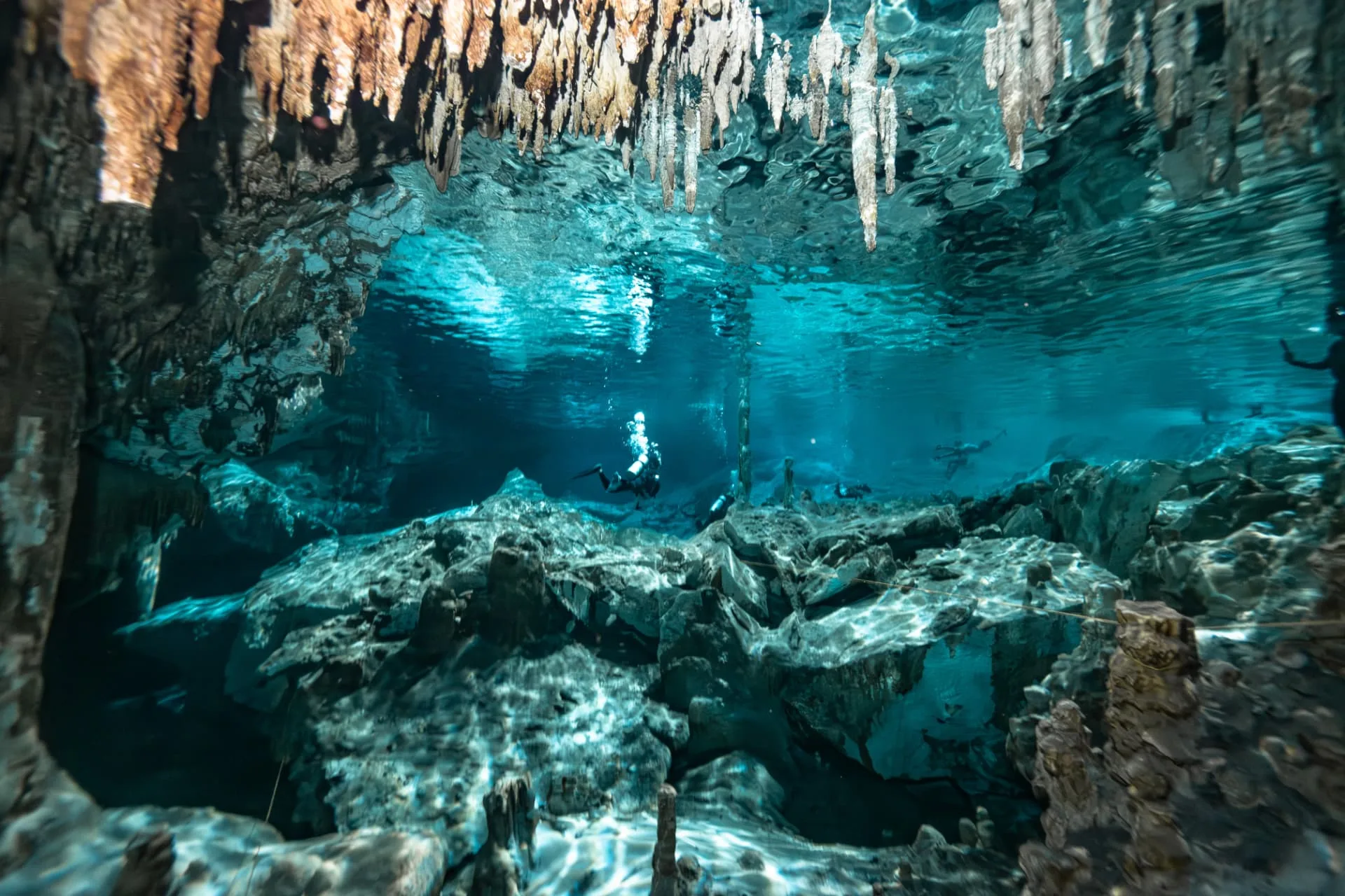 Stalactites hanging above clear water and light in Taak Bi Ha cenote, with a distant diver.