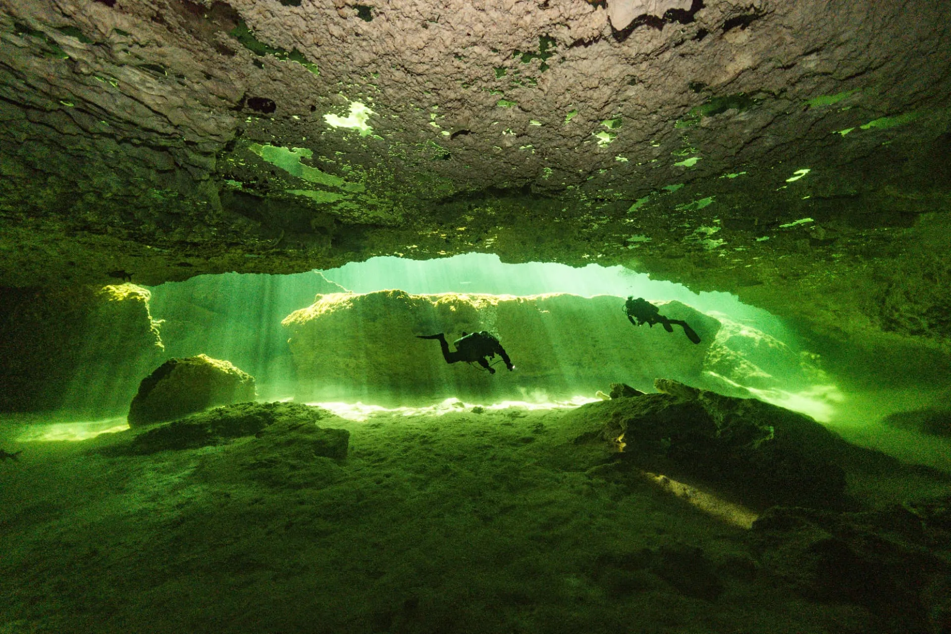 Two divers passing under a rock overhang lit green at Casa Cenote.