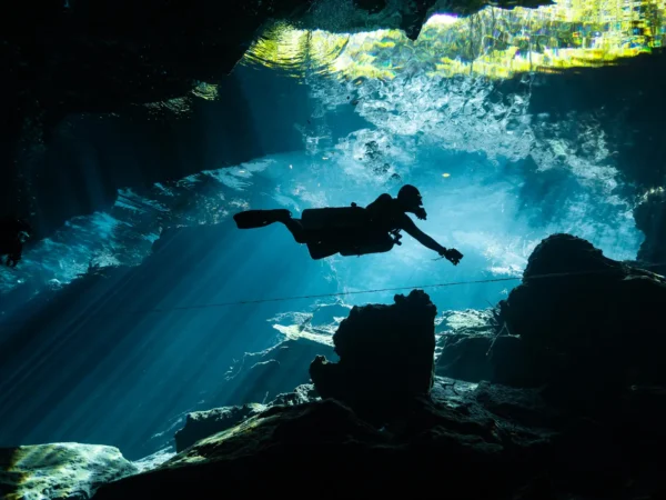 Diver exploring Cenote Kukulkán with light beams piercing the water and stalactites in the foreground – little brother kukulkan cenote dive