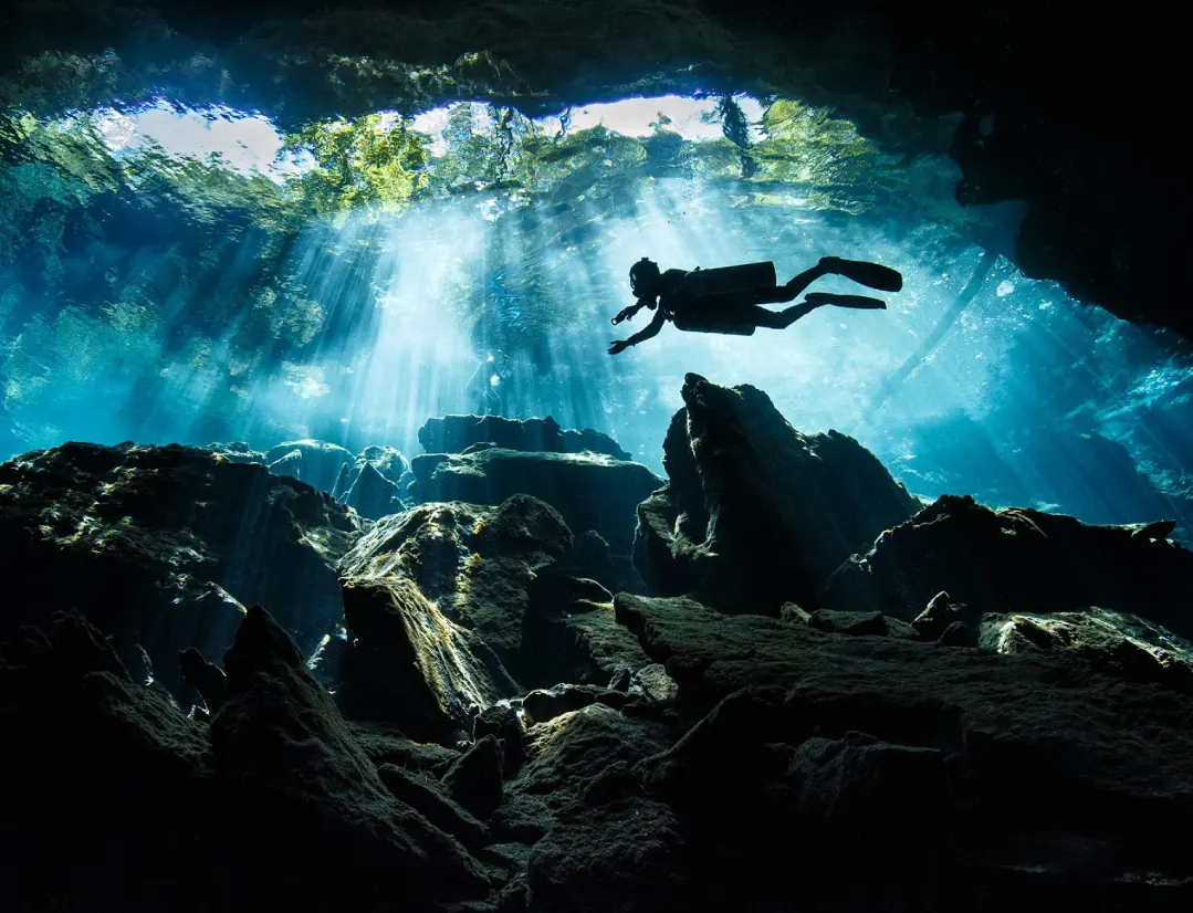 Buceador en caverna subacuática iluminada por haces de luz natural con formaciones rocosas y visibilidad cristalina